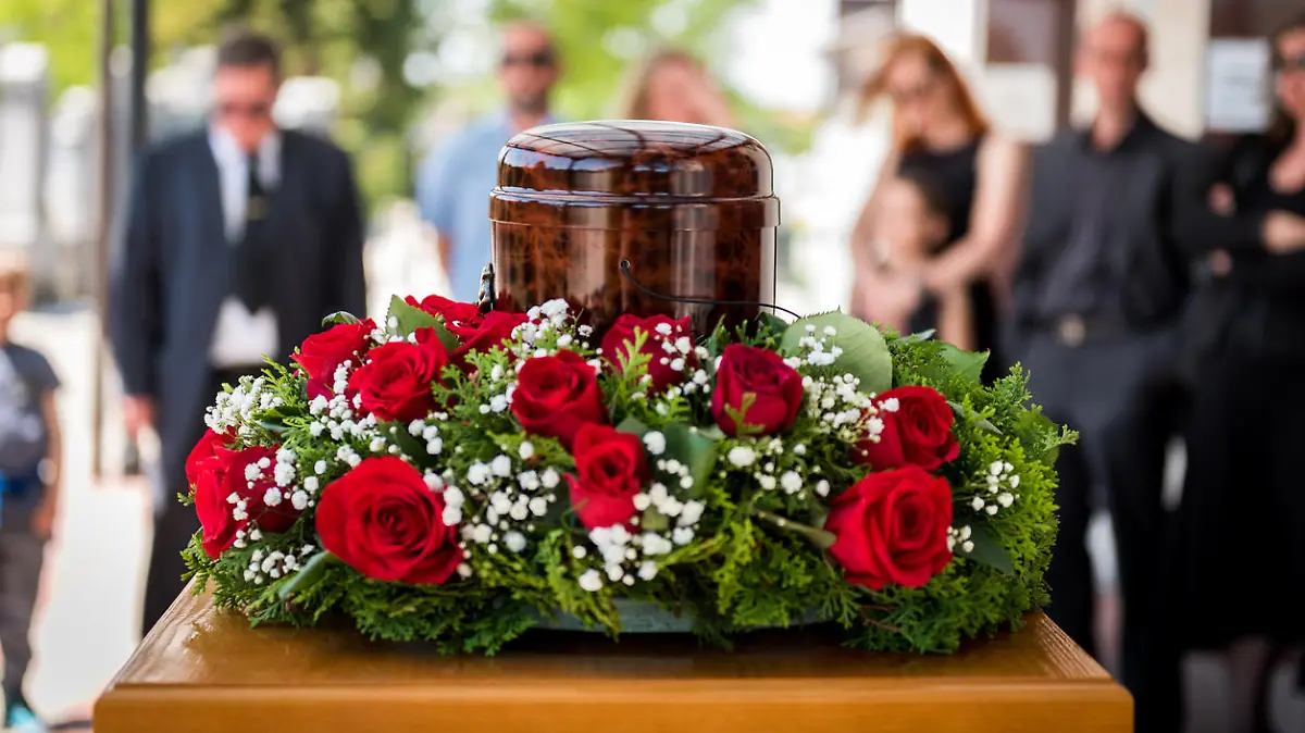 Funerary urn with ashes of dead and flowers at funeral. Burial urn decorated with flowers and people mourning in background at memorial service, sad and grieving last farewell to deceased person.