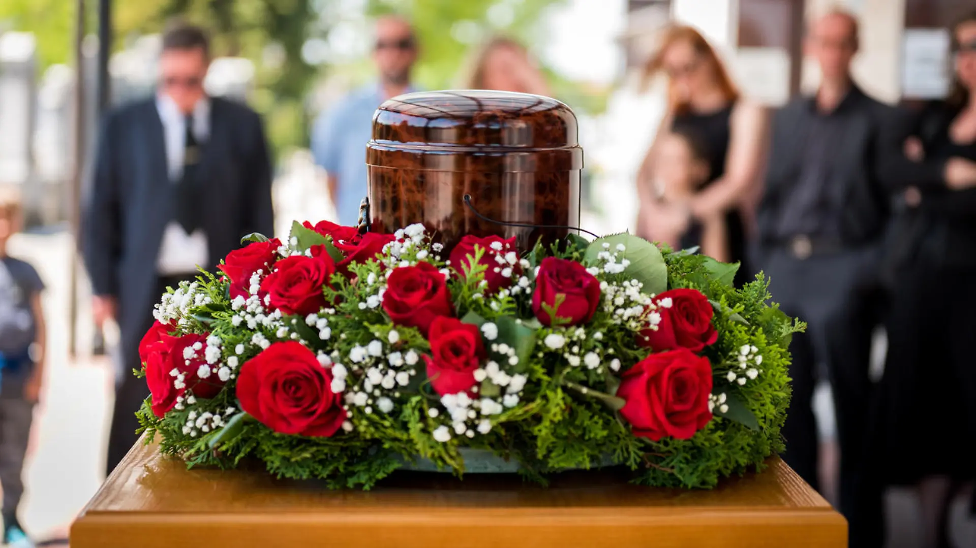 Funerary urn with ashes of dead and flowers at funeral. Burial urn decorated with flowers and people mourning in background at memorial service, sad and grieving last farewell to deceased person.