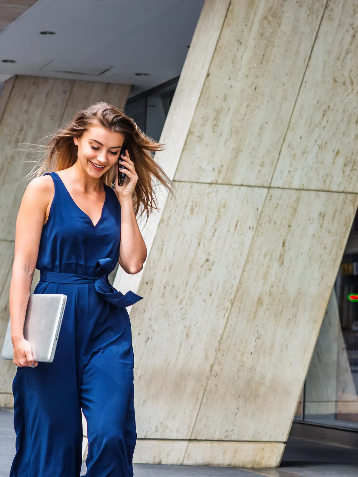 Young Eastern European American Woman talking on cell phone, traveling, working in New York City, wearing blue sleeveless jumpsuit, carrying laptop computer, walking on street outside office building."n
