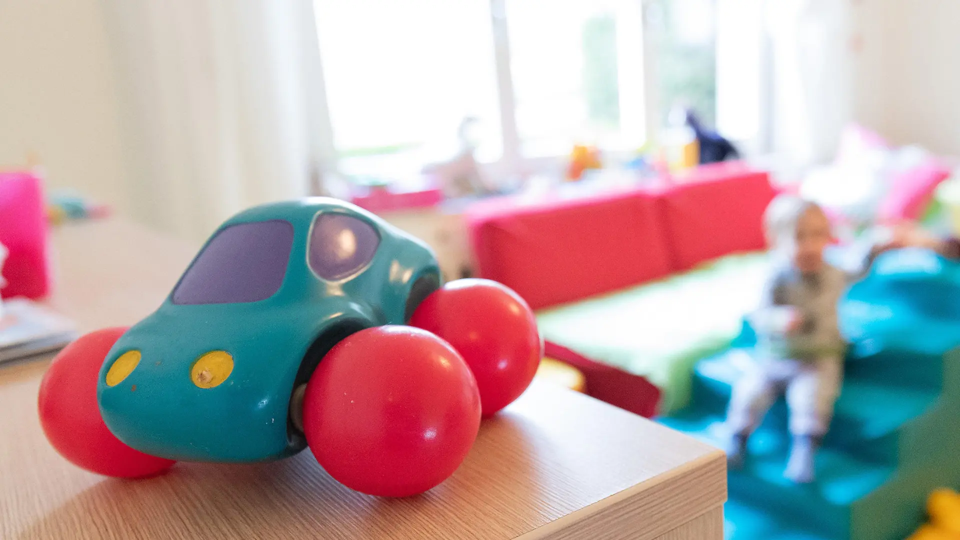 ARCHIV - 03.03.2020, Nordrhein-Westfalen, Bielefeld: ILLUSTRATION - Ein Spielzeugauto ist in den Räumen einer Kindertagesstätte zu sehen. Im Hintergrund ein Kind zusammen mit einer Erzieherin. Kinder in Nordrhein-Westfalen und anderen Bundesländern werden in der Corona-Krise nicht zu einem bundeseinheitlichen Stichtag in die Kitas zurückkehren. Das stellte NRW-Familienminister Stamp (FDP) am Montag klar. Foto: Friso Gentsch/dpa +++ dpa-Bildfunk +++