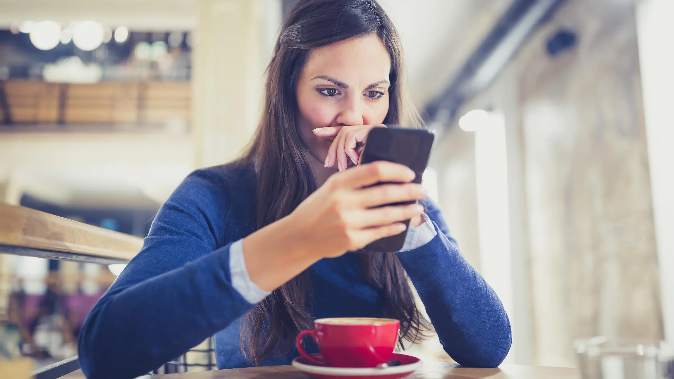 Young woman drink a cappuccino at the cafe, using mobile phone, texting. She is worried and suspicious