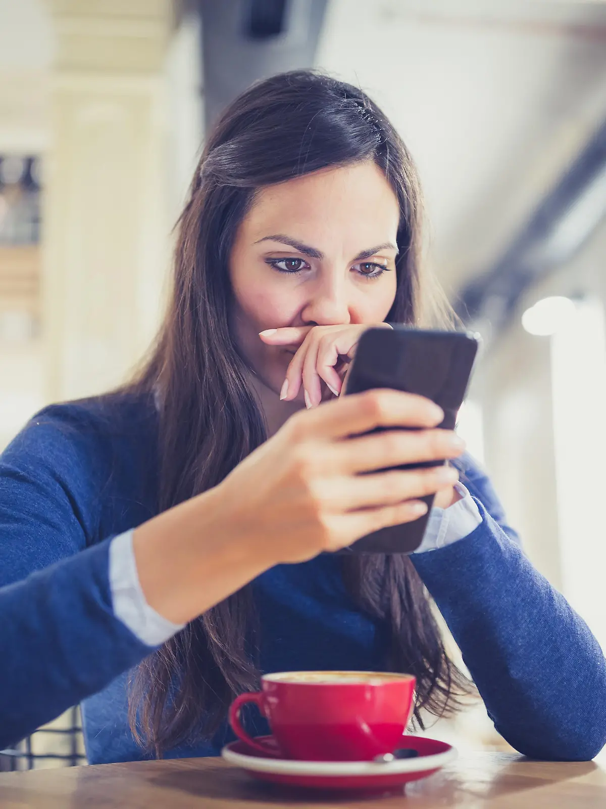 Young woman drink a cappuccino at the cafe, using mobile phone, texting. She is worried and suspicious