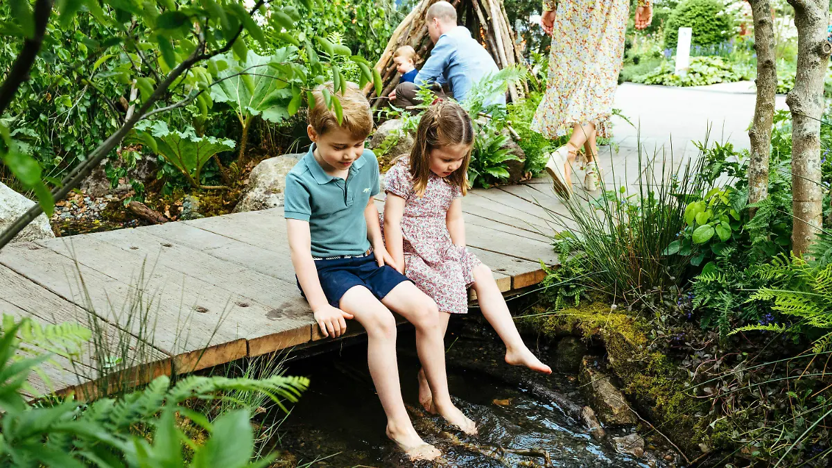 HANDOUT - 19.05.2019, Großbritannien, London: Der britische Prinz George und seine Schwester, Prinzessin Charlotte, sitzen auf einem Steg über einen Bach auf dem Gelände des von Adam White und Andree Davies mitgestalteten Garten vor der RHS Chelsea Flower Show, während im Hintergrund Prinz William (r) mit Prinz Louis zu sehen ist. Foto: Matt Porteous/Kensington Palace/AP/dpa - ACHTUNG: Nur zur redaktionellen Verwendung im Zusammenhang mit der aktuellen Berichterstattung und nur mit vollständiger Nennung des vorstehenden Credits +++ dpa-Bildfunk +++