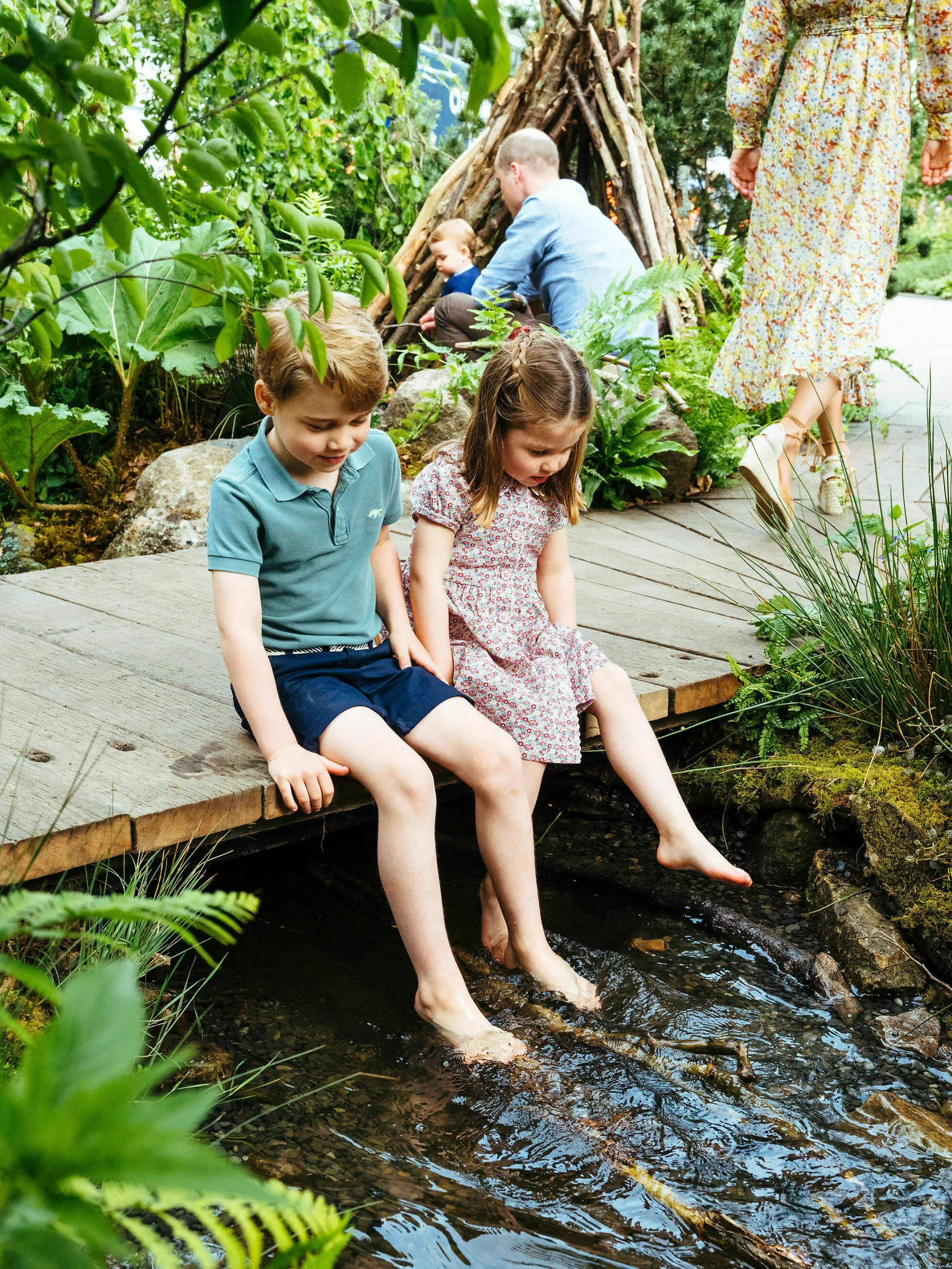 HANDOUT - 19.05.2019, Großbritannien, London: Der britische Prinz George und seine Schwester, Prinzessin Charlotte, sitzen auf einem Steg über einen Bach auf dem Gelände des von Adam White und Andree Davies mitgestalteten Garten vor der RHS Chelsea Flower Show, während im Hintergrund Prinz William (r) mit Prinz Louis zu sehen ist. Foto: Matt Porteous/Kensington Palace/AP/dpa - ACHTUNG: Nur zur redaktionellen Verwendung im Zusammenhang mit der aktuellen Berichterstattung und nur mit vollständiger Nennung des vorstehenden Credits +++ dpa-Bildfunk +++
