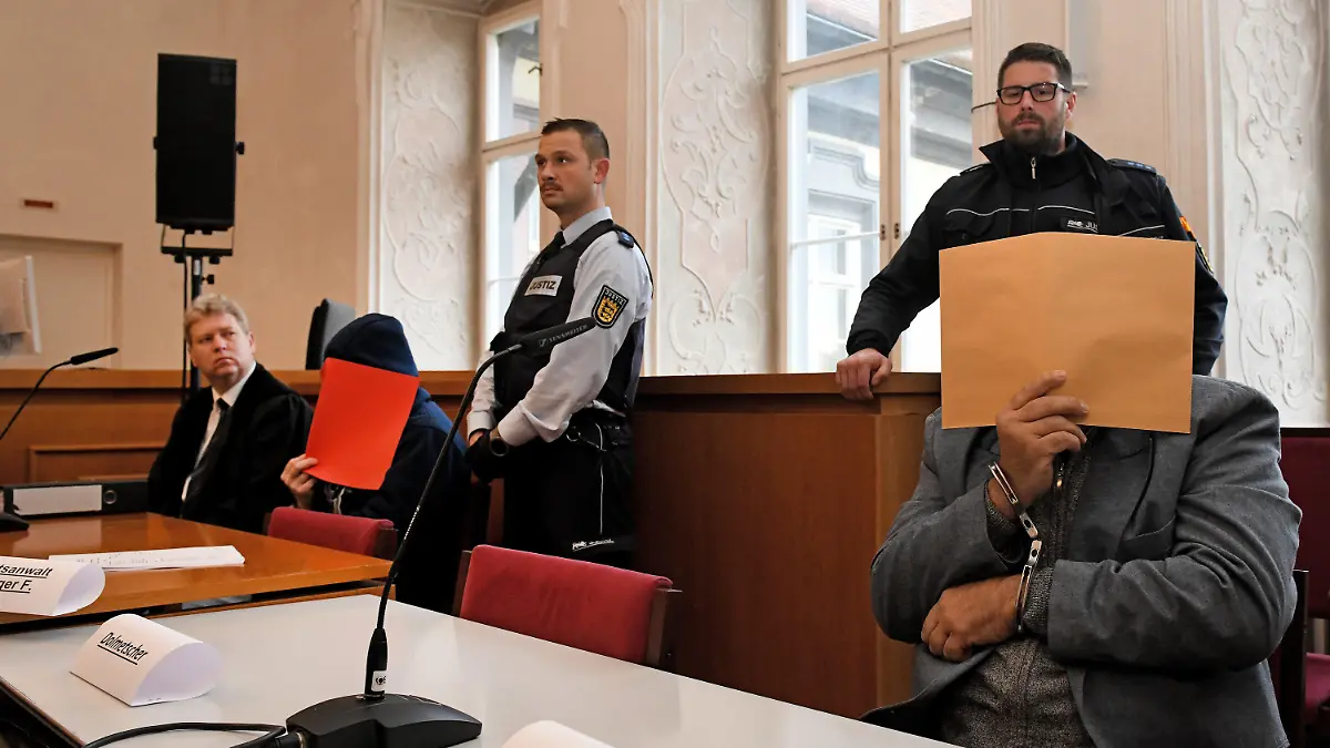 26.11.2019, Baden-Württemberg, Ellwangen: Der angeklagte Vater (r) sitzt mit gefesselten Händen auf der Anklagebank und hält sich einen Aktendeckel vor das Gesicht, links der Verteidiger Stephan Bauer mit einem der angeklagten Söhne (rote Akte). Er soll laut Anklage 2008 den damaligen Ehemann seiner Tochter in Sontheim an der Brenz erwürgt haben. 2014 soll er mit Hilfe seiner Söhne den damaligen Lebensgefährten der Tochter getötet haben. 2019 soll der Vater zusammen mit dem älteren Sohn zudem einen Mann aus Habgier getötet haben. Foto: Stefan Puchner/dpa +++ dpa-Bildfunk +++