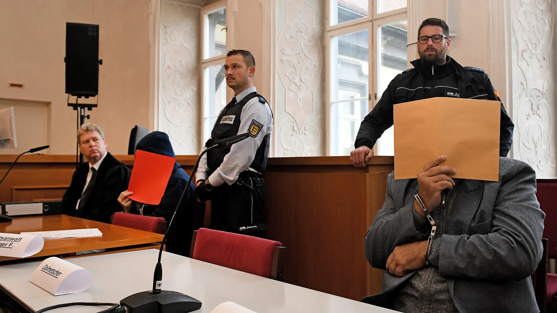 26.11.2019, Baden-Württemberg, Ellwangen: Der angeklagte Vater (r) sitzt mit gefesselten Händen auf der Anklagebank und hält sich einen Aktendeckel vor das Gesicht, links der Verteidiger Stephan Bauer mit einem der angeklagten Söhne (rote Akte). Er soll laut Anklage 2008 den damaligen Ehemann seiner Tochter in Sontheim an der Brenz erwürgt haben. 2014 soll er mit Hilfe seiner Söhne den damaligen Lebensgefährten der Tochter getötet haben. 2019 soll der Vater zusammen mit dem älteren Sohn zudem einen Mann aus Habgier getötet haben. Foto: Stefan Puchner/dpa +++ dpa-Bildfunk +++