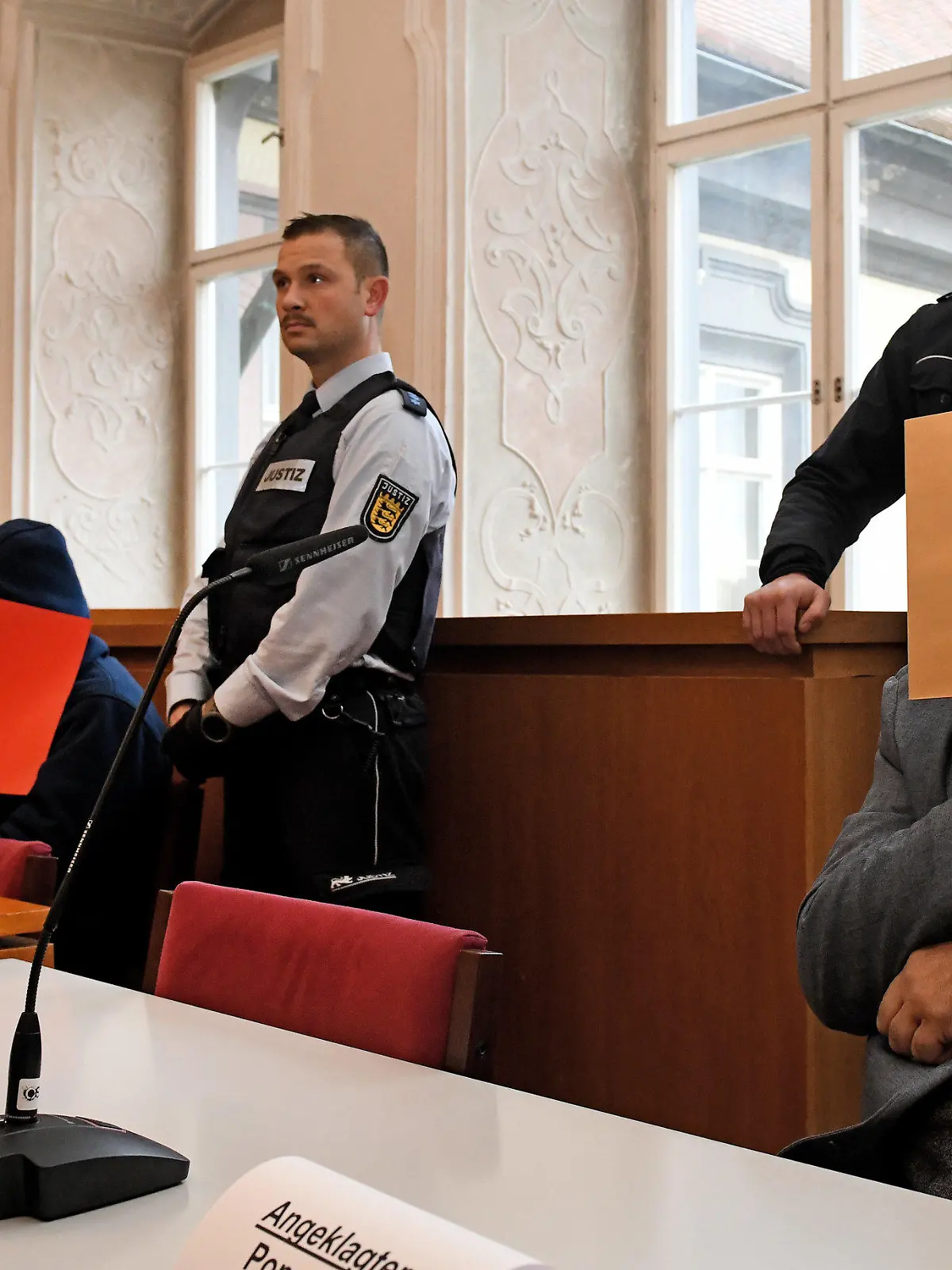 26.11.2019, Baden-Württemberg, Ellwangen: Der angeklagte Vater (r) sitzt mit gefesselten Händen auf der Anklagebank und hält sich einen Aktendeckel vor das Gesicht, links der Verteidiger Stephan Bauer mit einem der angeklagten Söhne (rote Akte). Er soll laut Anklage 2008 den damaligen Ehemann seiner Tochter in Sontheim an der Brenz erwürgt haben. 2014 soll er mit Hilfe seiner Söhne den damaligen Lebensgefährten der Tochter getötet haben. 2019 soll der Vater zusammen mit dem älteren Sohn zudem einen Mann aus Habgier getötet haben. Foto: Stefan Puchner/dpa +++ dpa-Bildfunk +++