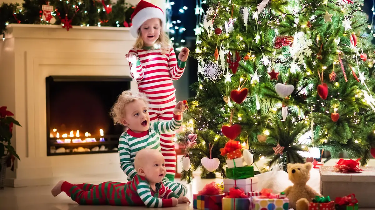Happy little kids in matching red and green striped pajamas decorate Christmas tree in beautiful living room with traditional fire place. Children opening presents on Xmas eve.