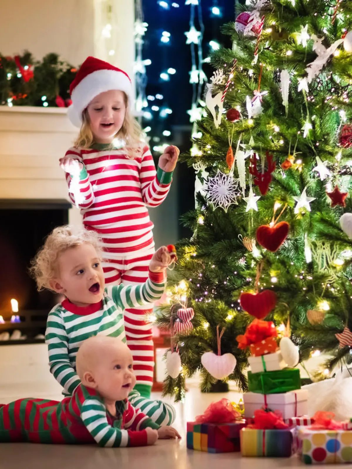 Happy little kids in matching red and green striped pajamas decorate Christmas tree in beautiful living room with traditional fire place. Children opening presents on Xmas eve.