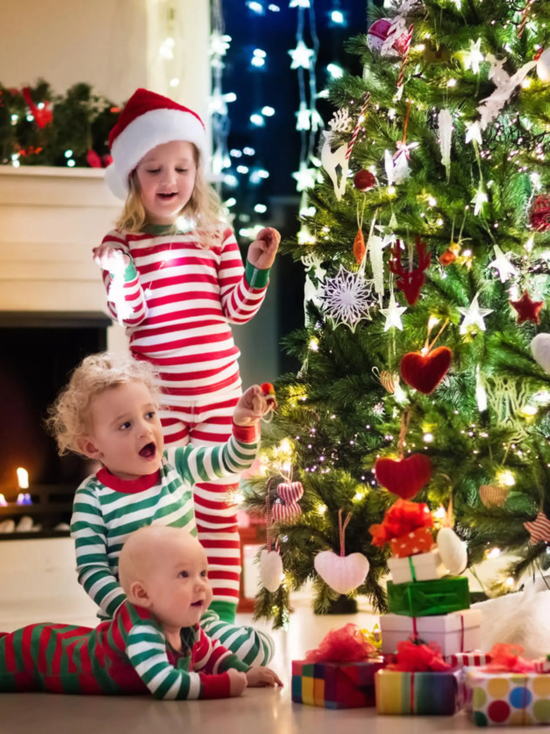 Happy little kids in matching red and green striped pajamas decorate Christmas tree in beautiful living room with traditional fire place. Children opening presents on Xmas eve.