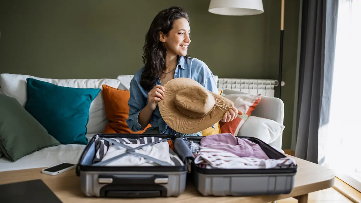 Young woman sitting on the couch and packing suitcase. She is holding a straw hat in her hands