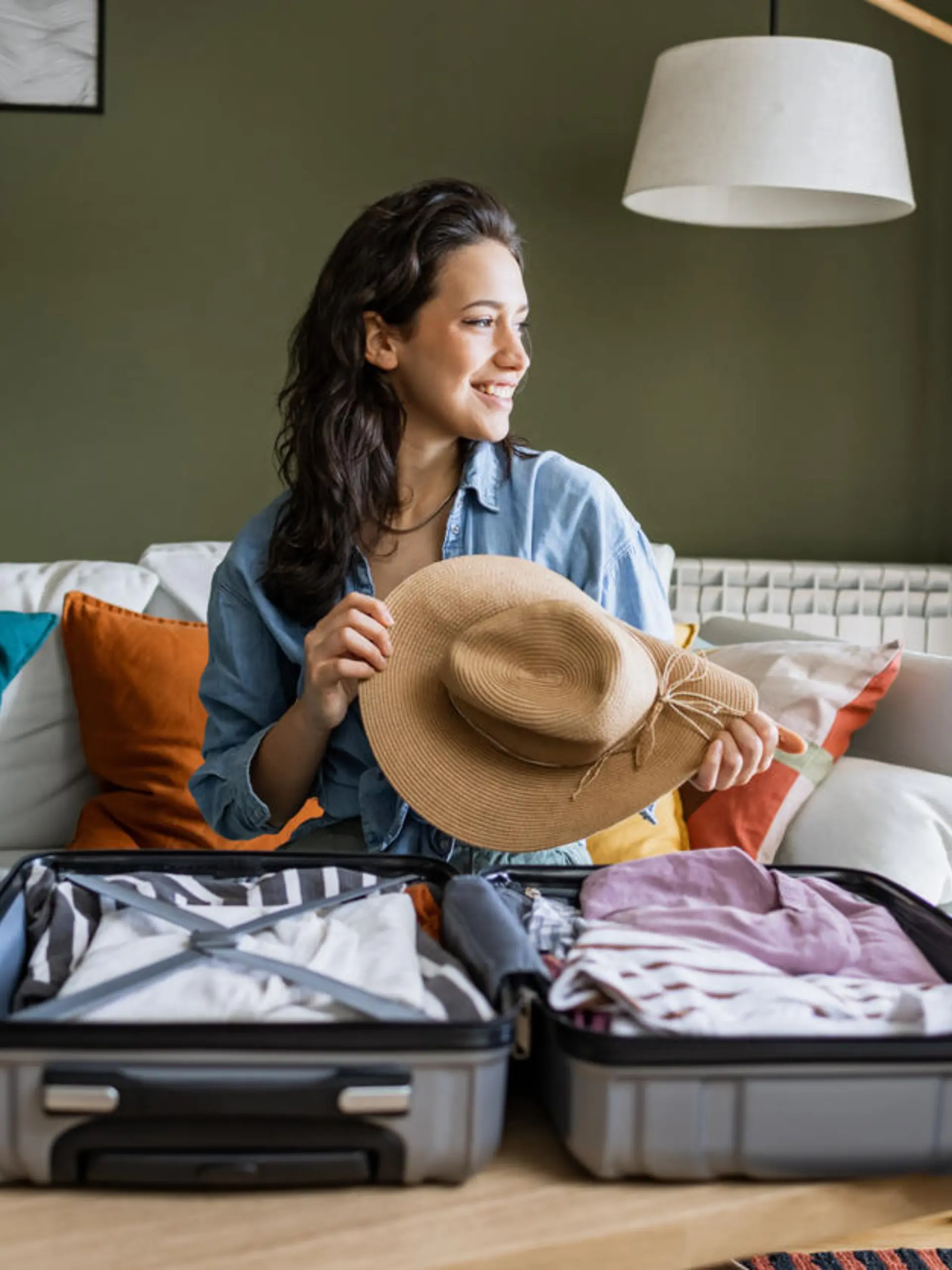 Young woman sitting on the couch and packing suitcase. She is holding a straw hat in her hands