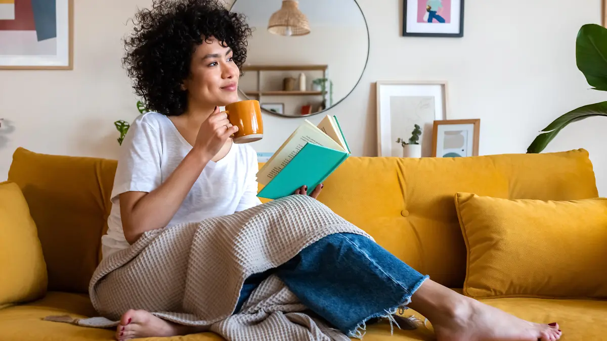 Pensive relaxed African american woman reading a book at home, drinking coffee sitting on the couch. Copy space. Lifestyle concept.