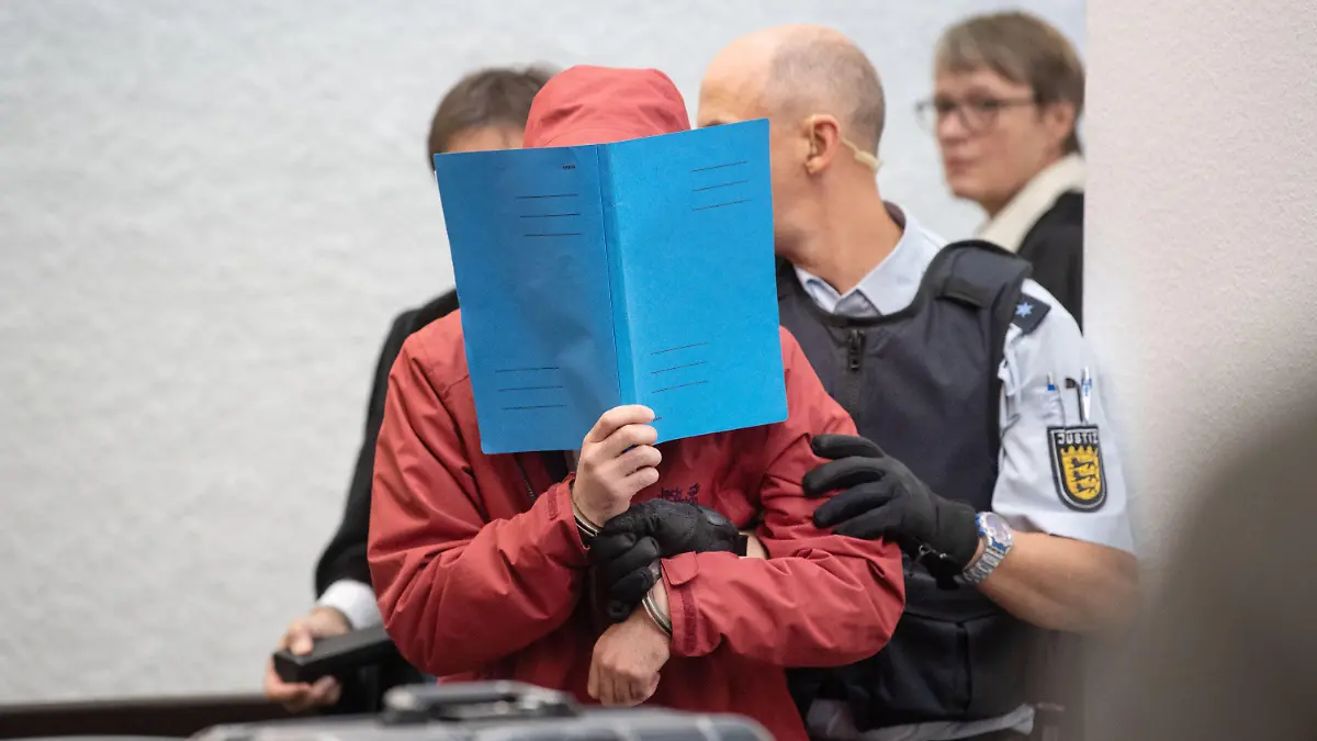 ARCHIV - 11.09.2019, Baden-Württemberg, Stuttgart: Der Angeklagte in einem Mordprozess nach einem tödlichem Unfall wird von einem Justizbeamten in einen Gerichtssaal des Landgerichts Stuttgart geführt. Im Hintergrund steht die Vorsitzende Richterin am Landgericht, Cornelie Eßlinger-Graf. Der Mann soll mit einem Mietwagen mit hohem Tempo unterwegs gewesen sein und prallte in einen Kleinwagen. Dessen 25 Jahre alter Fahrer und die 22 Jahre alte Beifahrerin starben. Es ist der erste Mordprozess gegen einen Raser in Baden-Württemberg. (zu dpa: «Im Mordprozess gegen einen Autofahrer nach einem tödlichen Unfall mit einem Sportwagen wird eine Aussage des mutmaßlichen Rasers erwartet») Foto: Marijan Murat/dpa +++ dpa-Bildfunk +++