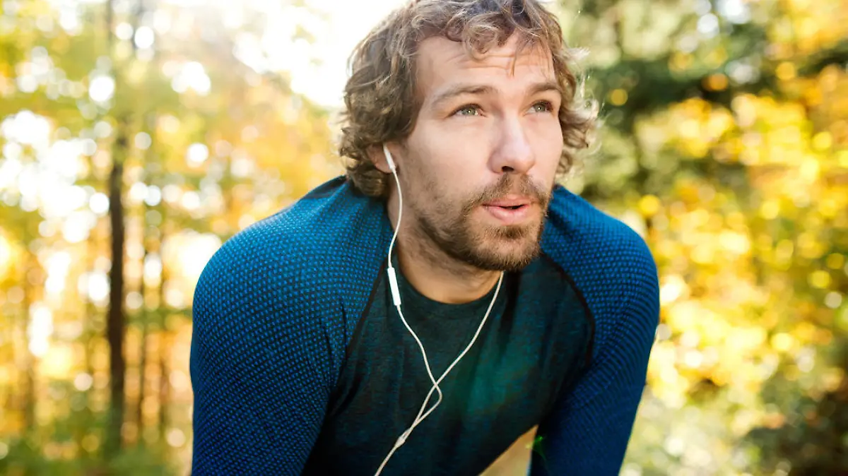 Young handsome runner with earphones in his ears, listening music, outside in sunny autumn nature, resting, breathing out
