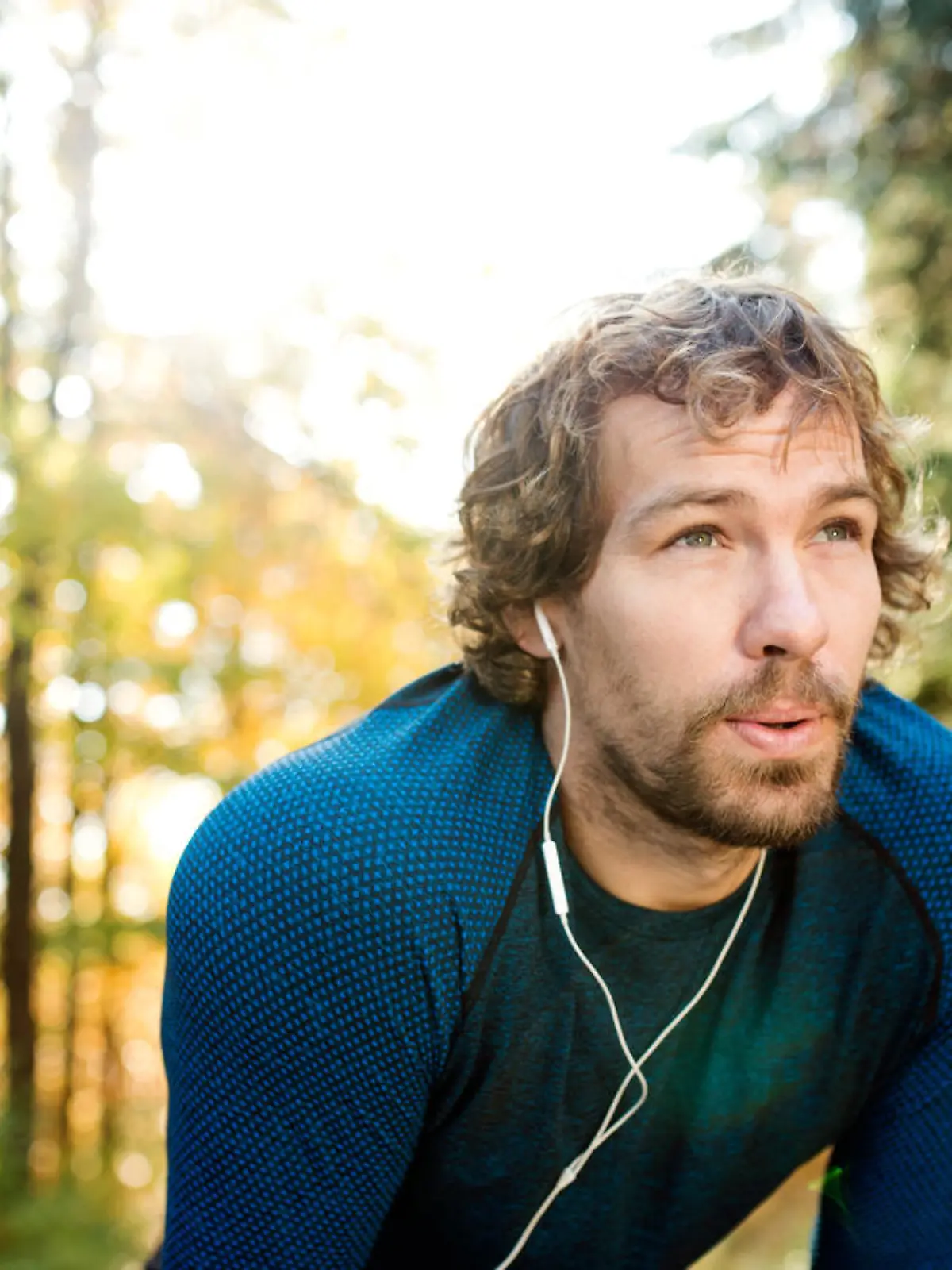 Young handsome runner with earphones in his ears, listening music, outside in sunny autumn nature, resting, breathing out