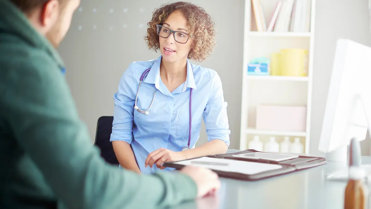 A female doctor sits at her desk and chats to a male patient about his illness . She is dressed in blouse with rolled up sleeves . She is explaining the upcoming procedure.