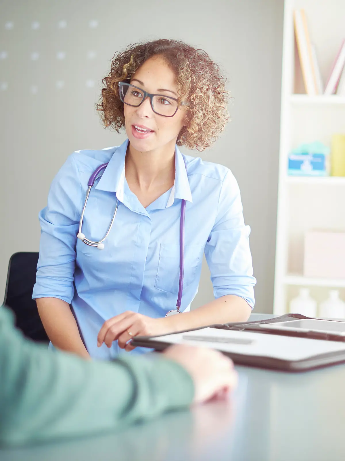 A female doctor sits at her desk and chats to a male patient about his illness . She is dressed in blouse with rolled up sleeves . She is explaining the upcoming procedure.