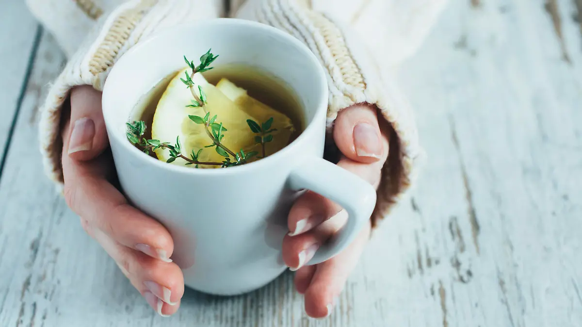 Human hands holding cup of tea with thyme herb and lemon slices on a wooden table,.