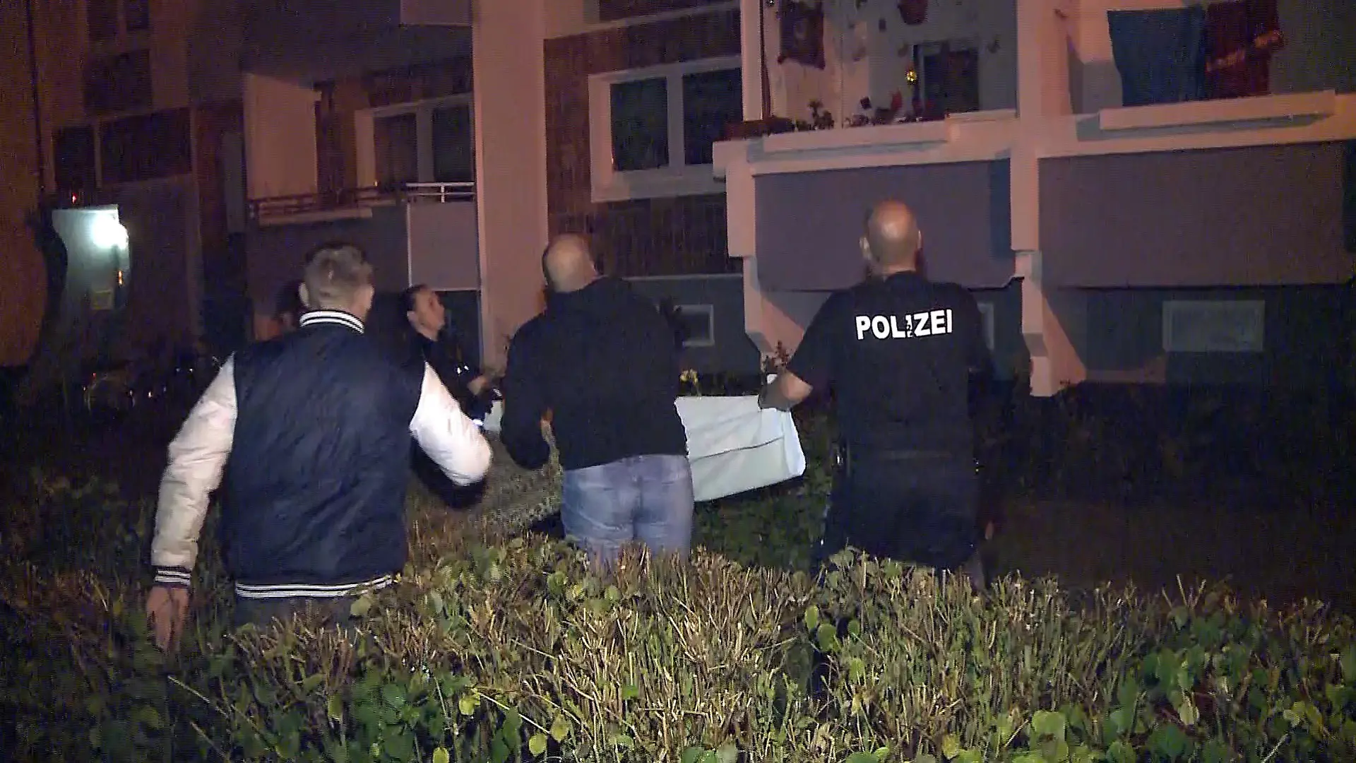 21.07.2019, Mecklenburg-Vorpommern, Rostock: Polizisten und Anwohner stehen vor einem Hochhaus und breiten ein Tuch aus, weil ein Mann in einem Obergeschoss einen Säugling über die Balkonbrüstung hält und droht, in fallen zu lassen. Der Mann hat mit einem Messer auf einen Säugling eingestochen und dem Baby dabei schwerste Verletzungen zugefügt. Anschließend sei der 40-Jährige über die Brüstung des Balkons der Wohnung im fünften Obergeschoss gestiegen und in die Tiefe gesprungen, sagte ein Polizeisprecher am Sonntag. Dabei sei er ums Leben gekommen. Das Baby wurde in der Universitätsklinik Rostock notoperiert. (Bestmögliche verfügbare Qualität) Foto: Stefan Tretropp/dpa +++ dpa-Bildfunk +++