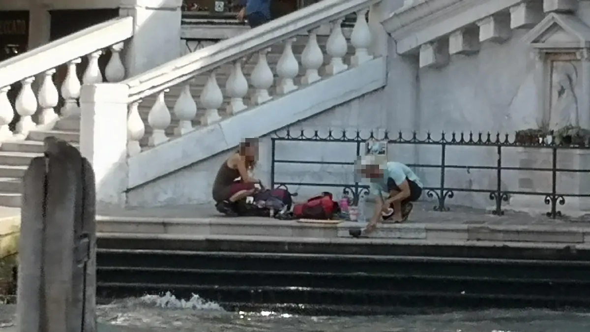 HANDOUT - 19.07.2019, Italien, Venedig: Zwei deutsche Touristen sitzen mit ihrem Gepäck an der Treppe der Rialto-Brücke und kochen sich mit einem kleinen Kaffeekocher einen Kaffee auf dem Gehweg. (zu dpa "Deutsche Touristen kochen Kaffee in Venedig - fast 1000 Euro Strafe") Foto: Comune di Venezia/dpa - ACHTUNG: Nur zur redaktionellen Verwendung im Zusammenhang mit der aktuellen Berichterstattung und nur mit vollständiger Nennung des vorstehenden Credits +++ dpa-Bildfunk +++