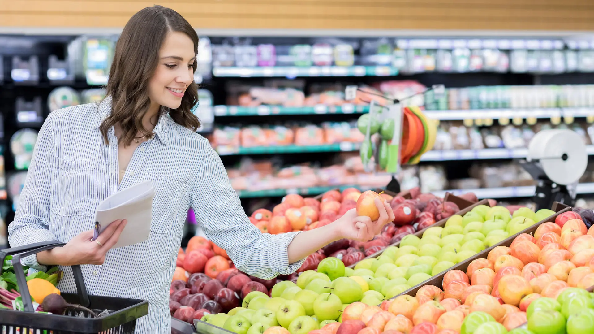 A cheerful young woman stands in the produce section of a grocery store. She has a shopping basket on her arm as she carefully inspects an apple.