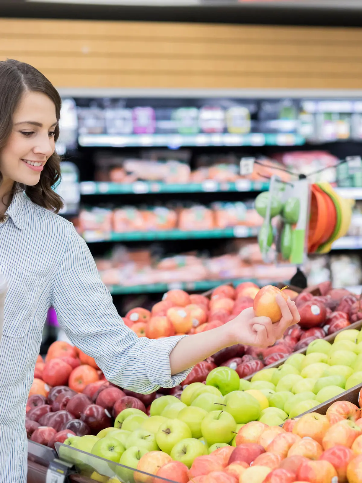 A cheerful young woman stands in the produce section of a grocery store.  She has a shopping basket on her arm as she carefully inspects an apple.