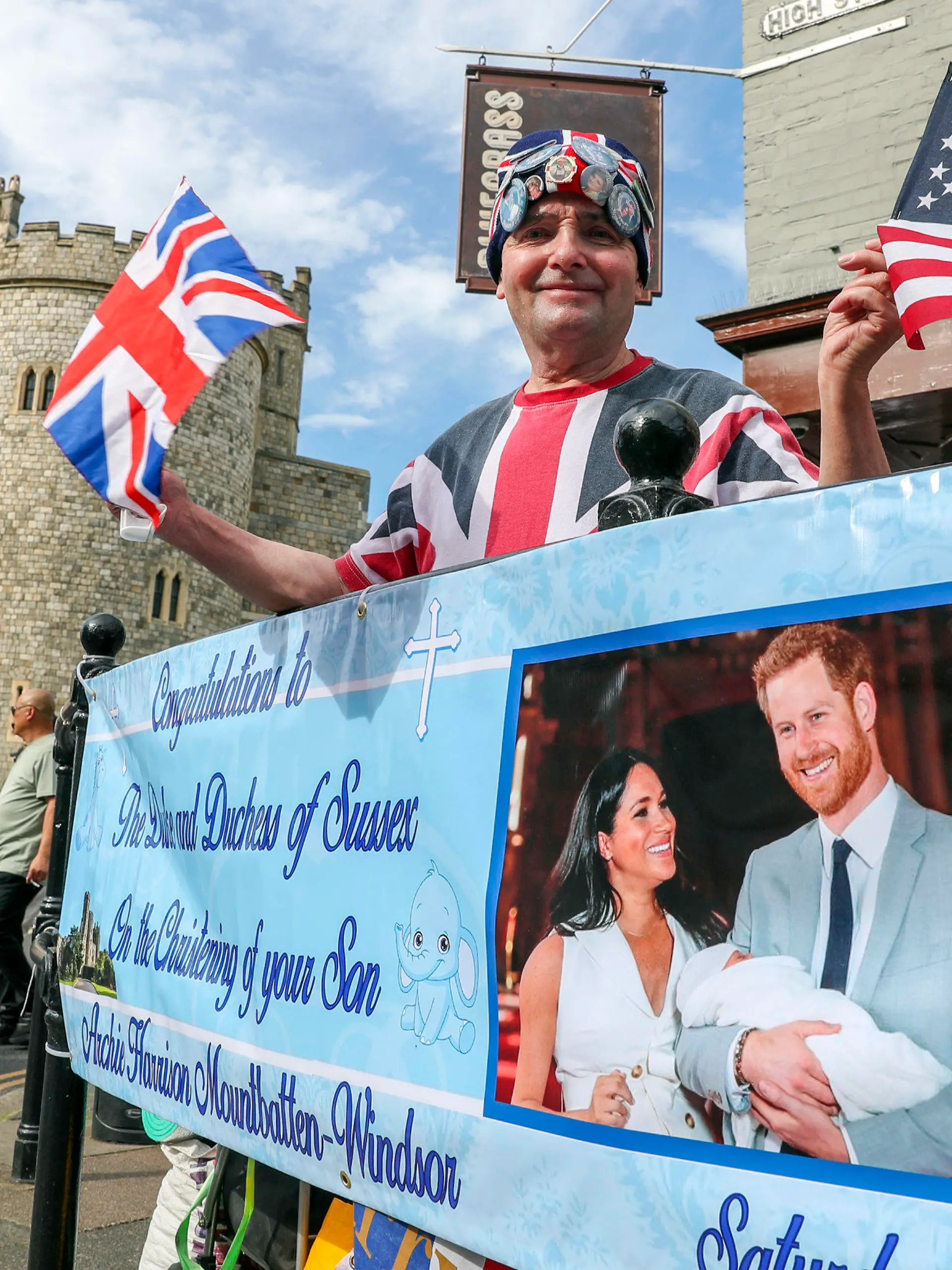 05.07.2019, Großbritannien, Windsor: John Loughrey, Fan der britischen Königsfamilie, steht hinter einem Banner, das ein Bild von Prinz Harry und Herzogin Meghan mit ihrem Sohn Archie zeigt. Archie wird am Samstag, den 05.07.2019, in Windsor getauft. Die Zeremonie soll in einem kleinen Kreis stattfinden. Foto: Steve Parsons/PA Wire/dpa +++ dpa-Bildfunk +++