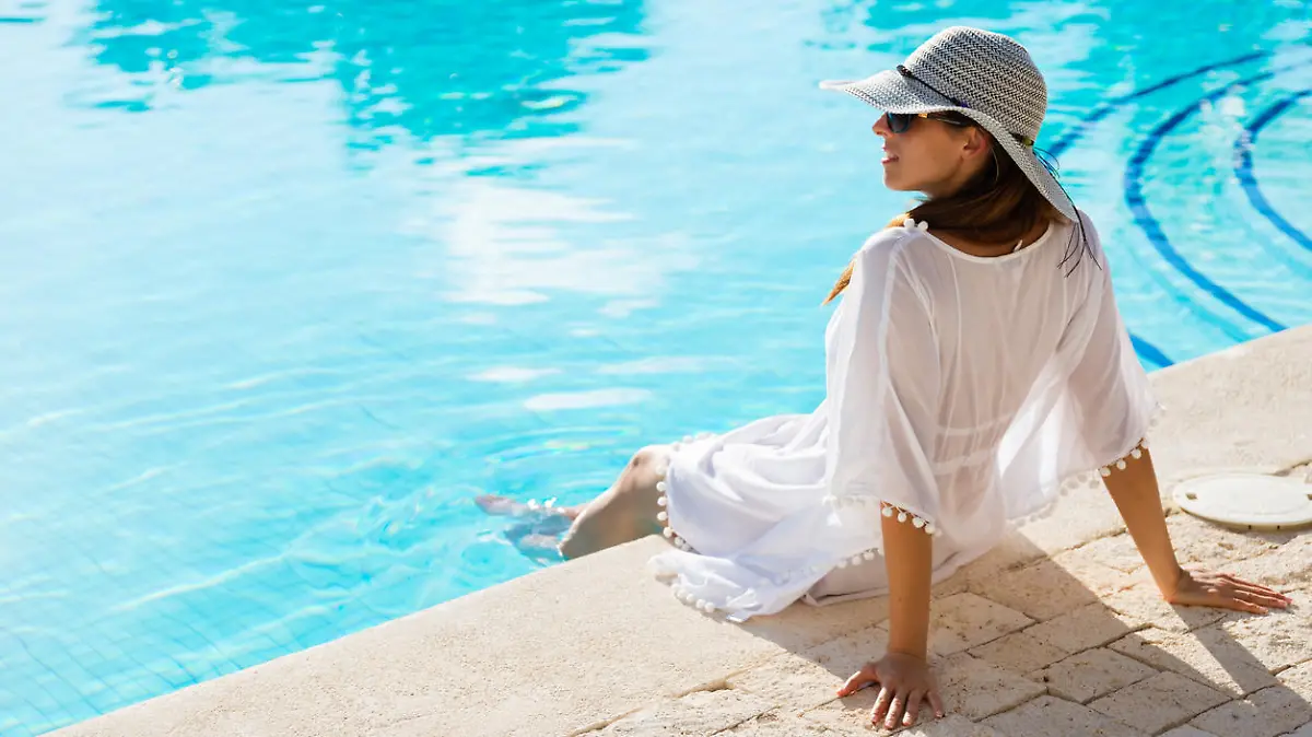Fashion beautiful woman on summer vacation relaxing at luxury resort spa poolside. Young fashionable lady wearing sun hat and white kaftan.