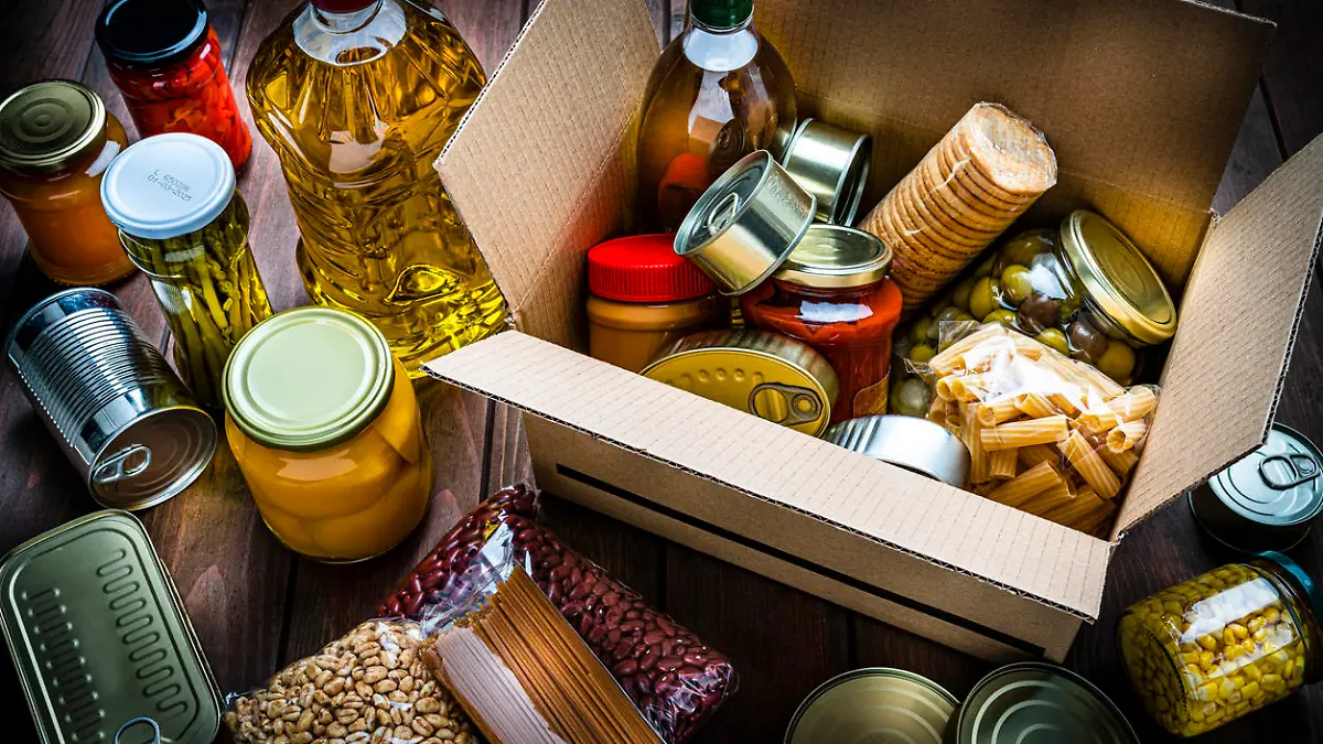 High angle view of a cardboard box filled with multicolored non-perishable canned goods, conserves, sauces and oils shot on wooden table. The composition includes cooking oil bottle, pasta, crackers, preserves and tins. High resolution 42Mp studio digital capture taken with SONY A7rII and Zeiss Batis 40mm F2.0 CF lens