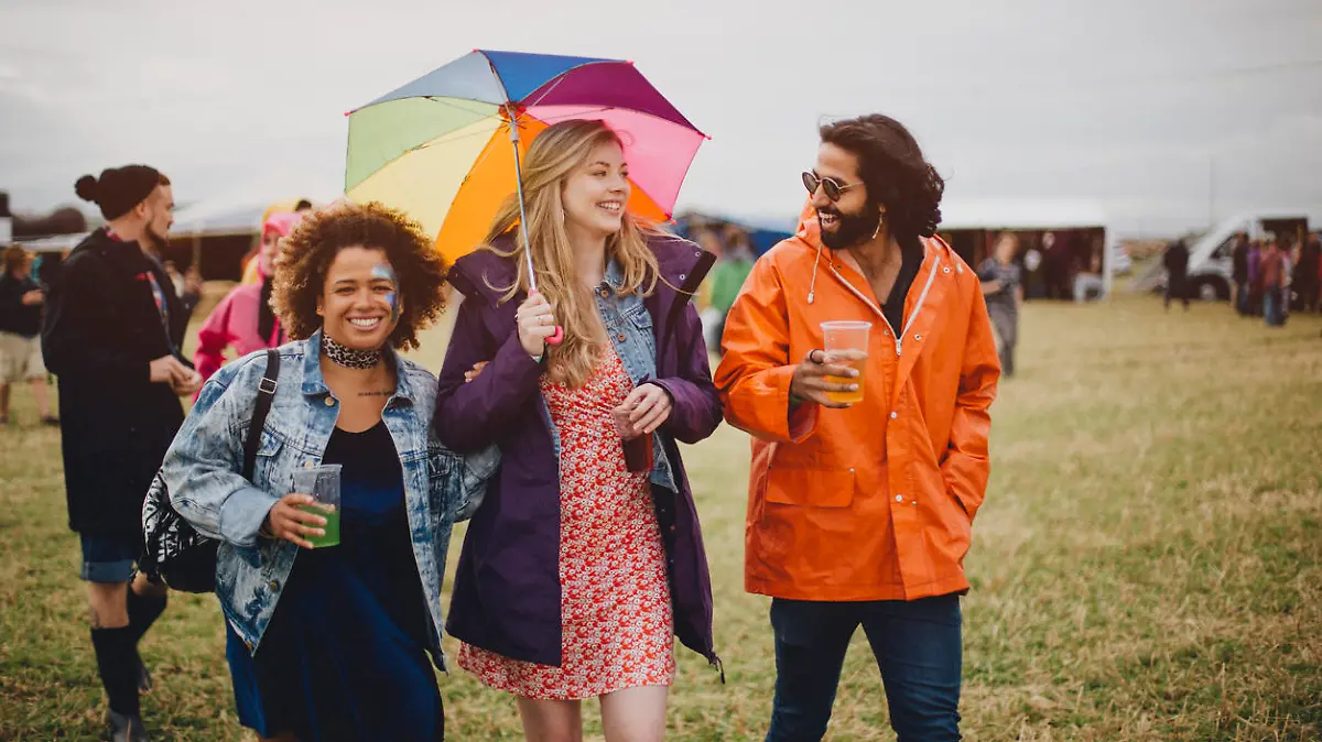 Group of friends are walking together to go and watch a live performance at a music festival. It is raining and they have an umbrella and raincoats. They are enjoying some alcoholic drinks.