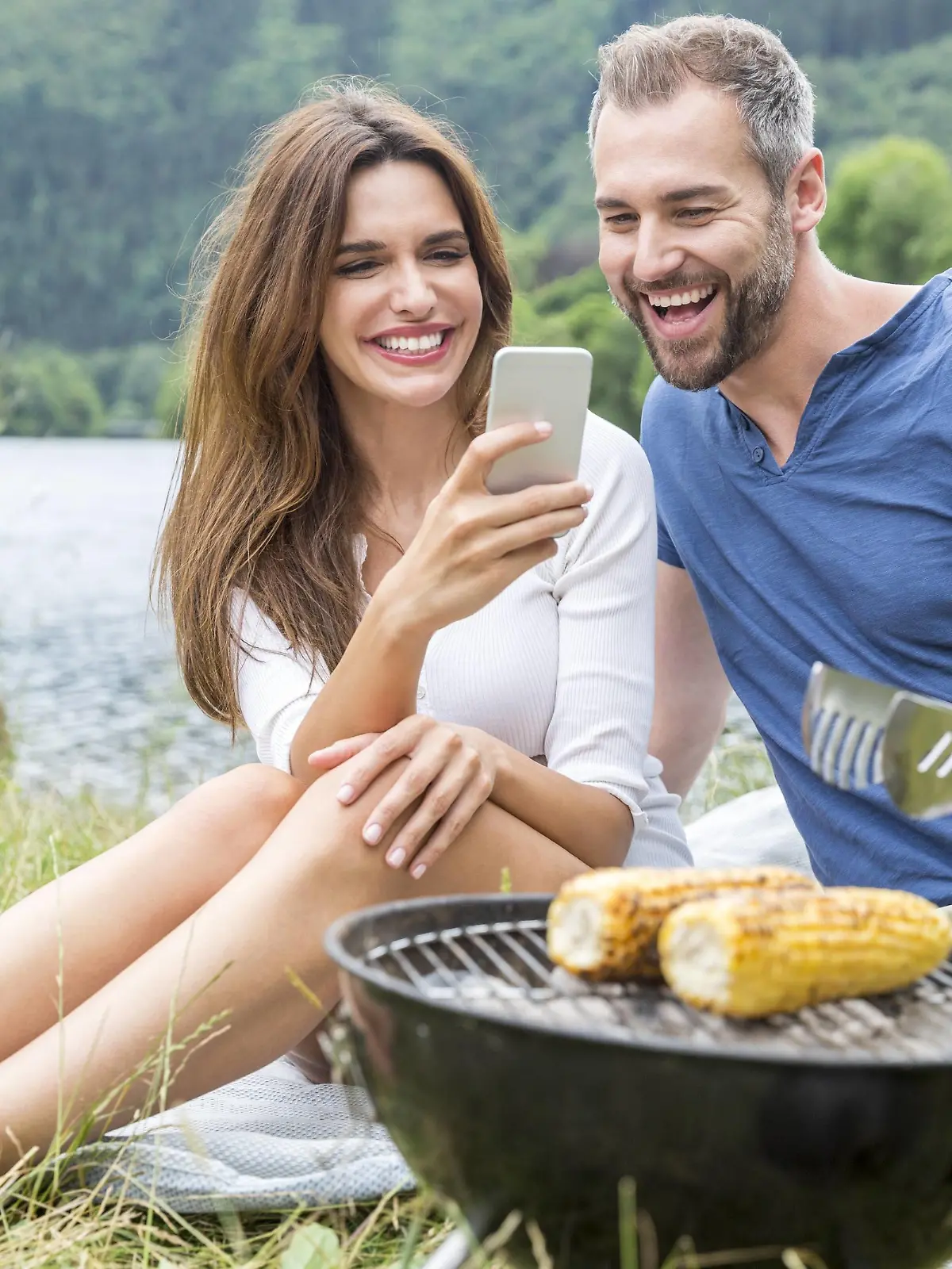 Happy couple with cell phone having a barbecue at lakeside model released Symbolfoto PUBLICATIONxINxGERxSUIxAUTxHUNxONLY FMKF002847  
