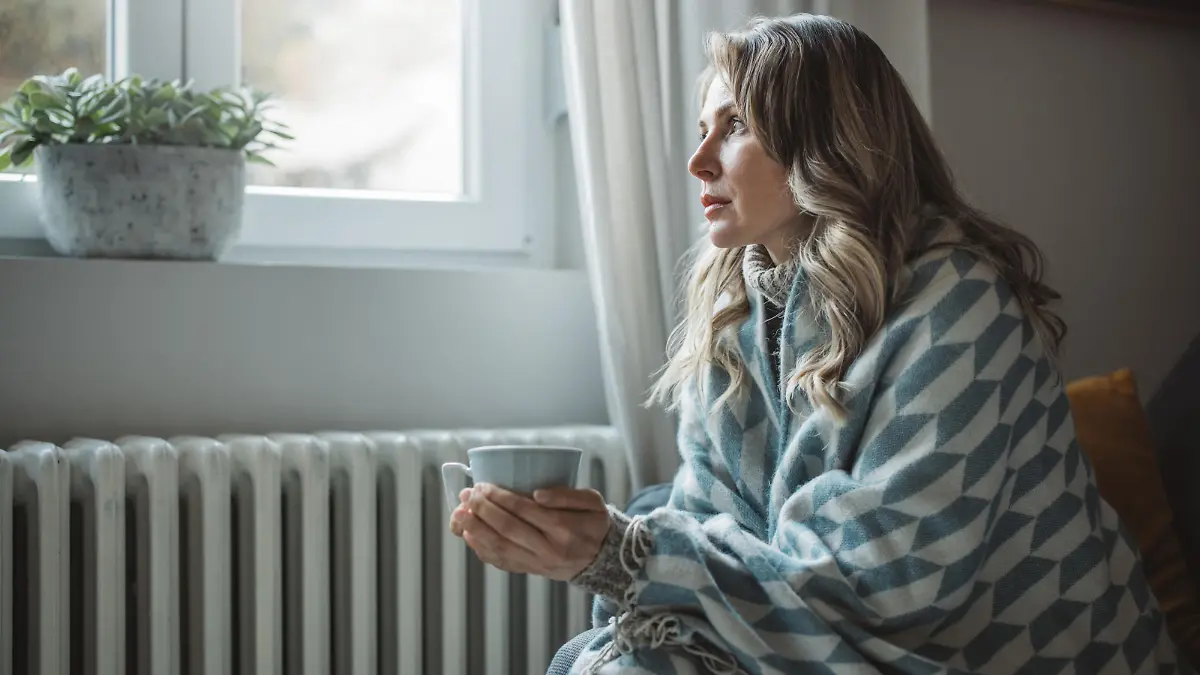 Unwell mature woman in blanket sit in cold living room suffer from air conditioner lack. She is sitting next to radiator.No heating concept.