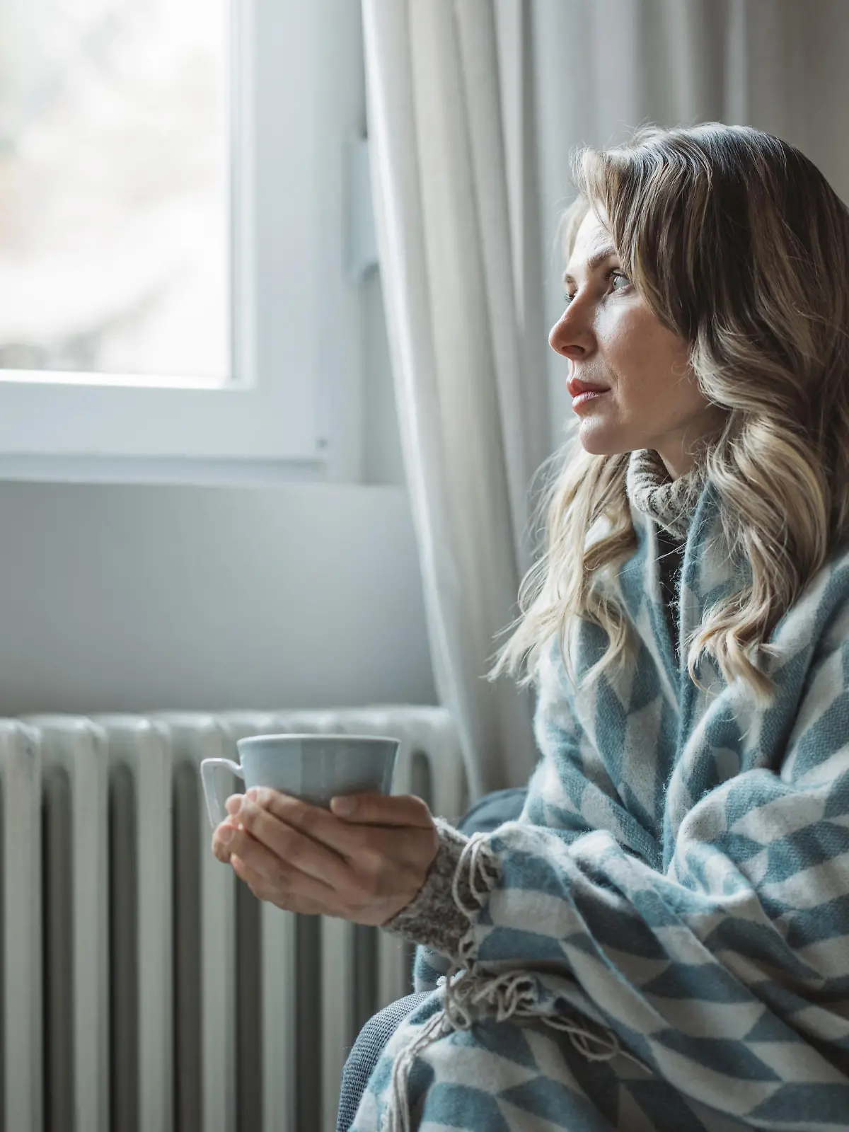 Unwell mature woman in blanket sit in cold living room suffer from air conditioner lack. She is sitting next to radiator.No heating concept.