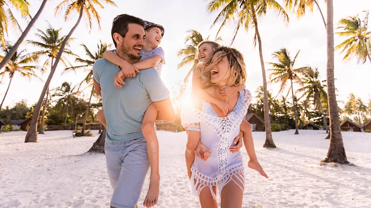 Happy parents and their small kids having fun while piggybacking on the beach in summer day.