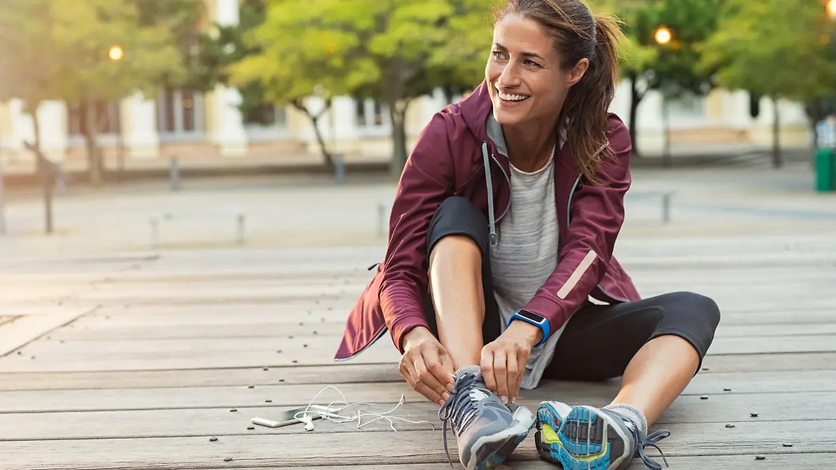 Mature fitness woman tie shoelaces on road. Cheerful runner sitting on floor on city streets with mobile and earphones wearing sport shoes. Active latin woman tying shoe lace before running.