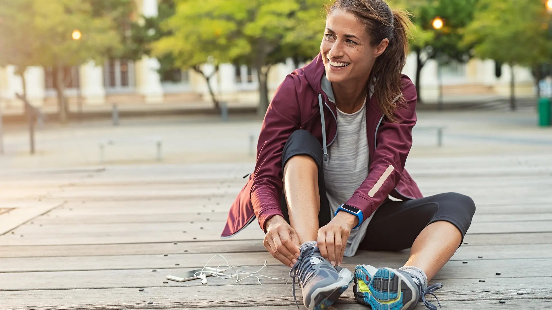 Mature fitness woman tie shoelaces on road. Cheerful runner sitting on floor on city streets with mobile and earphones wearing sport shoes. Active latin woman tying shoe lace before running.