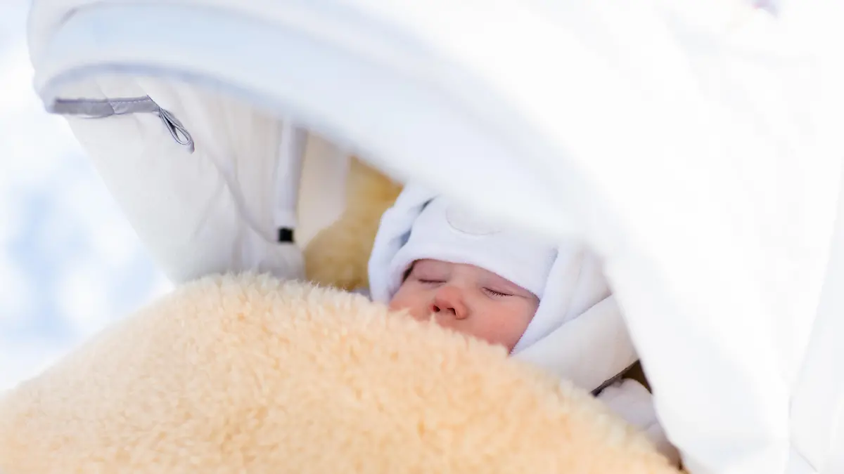 Cute newborn child sleeping in stroller on a cold winter day. New born baby taking a nap in warm sheepskin fur footmuff in a stroller in snowy park. Kids sleep outdoors in pram. Family fun in snow.