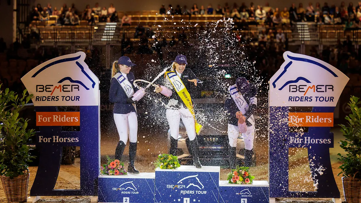 19.02.2023, Schleswig-Holstein, Neumünster: Pferdesport/Springreiten: Großer Preis (Finale der Riders Tour). Janne-Friederike Meyer-Zimmermann (M) wird bei der Siegerehrung von Sophie Hinners (l) und Mathias Norheden Johansen mit einer Sektdusche gefeiert. Meyer-Zimmermann hat die Gesamtwertung der Riders Tours gewonnen. (zu «Meyer-Zimmermann Gesamtsiegerin der Riders Tour») Foto: Stefan Lafrentz/dpa +++ dpa-Bildfunk +++