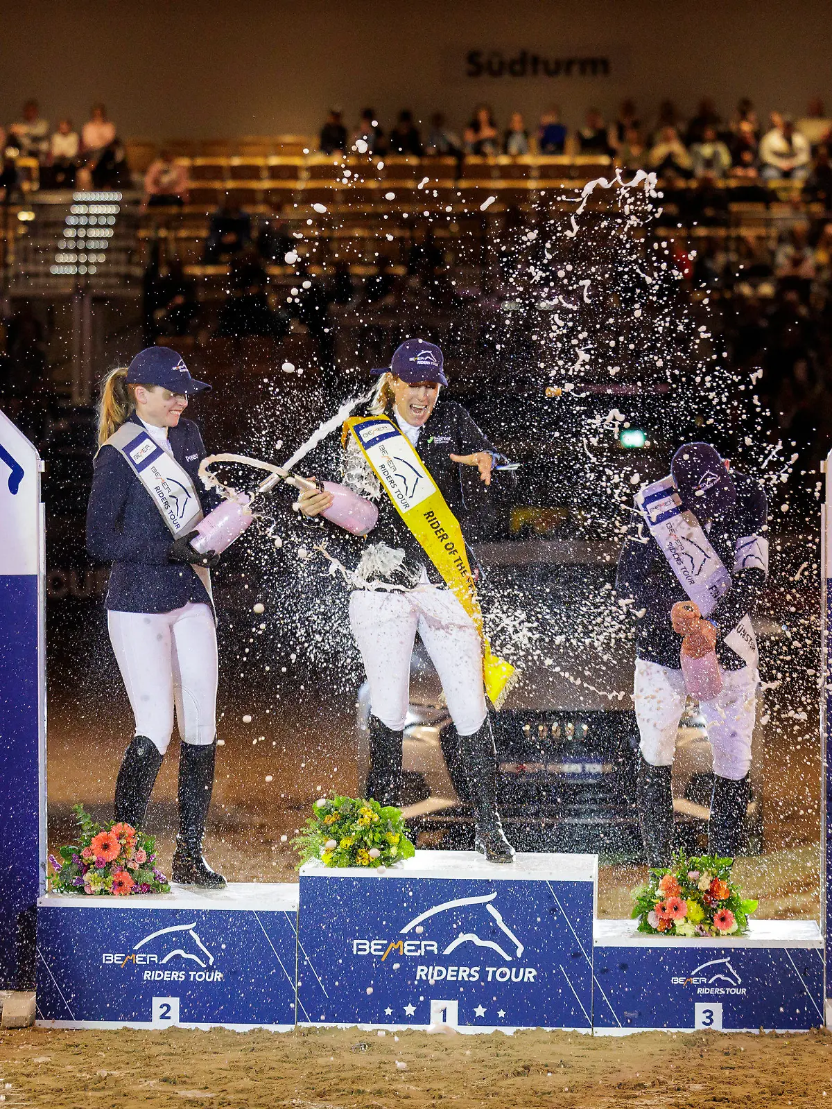 19.02.2023, Schleswig-Holstein, Neumünster: Pferdesport/Springreiten: Großer Preis (Finale der Riders Tour). Janne-Friederike Meyer-Zimmermann (M) wird bei der Siegerehrung von Sophie Hinners (l) und Mathias Norheden Johansen mit einer Sektdusche gefeiert. Meyer-Zimmermann hat die Gesamtwertung der Riders Tours gewonnen. (zu «Meyer-Zimmermann Gesamtsiegerin der Riders Tour») Foto: Stefan Lafrentz/dpa +++ dpa-Bildfunk +++