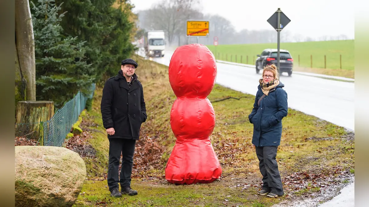 Neben einer ländlichen Straße steht eine große rote Kunstfigur zwischen zwei Menschen.