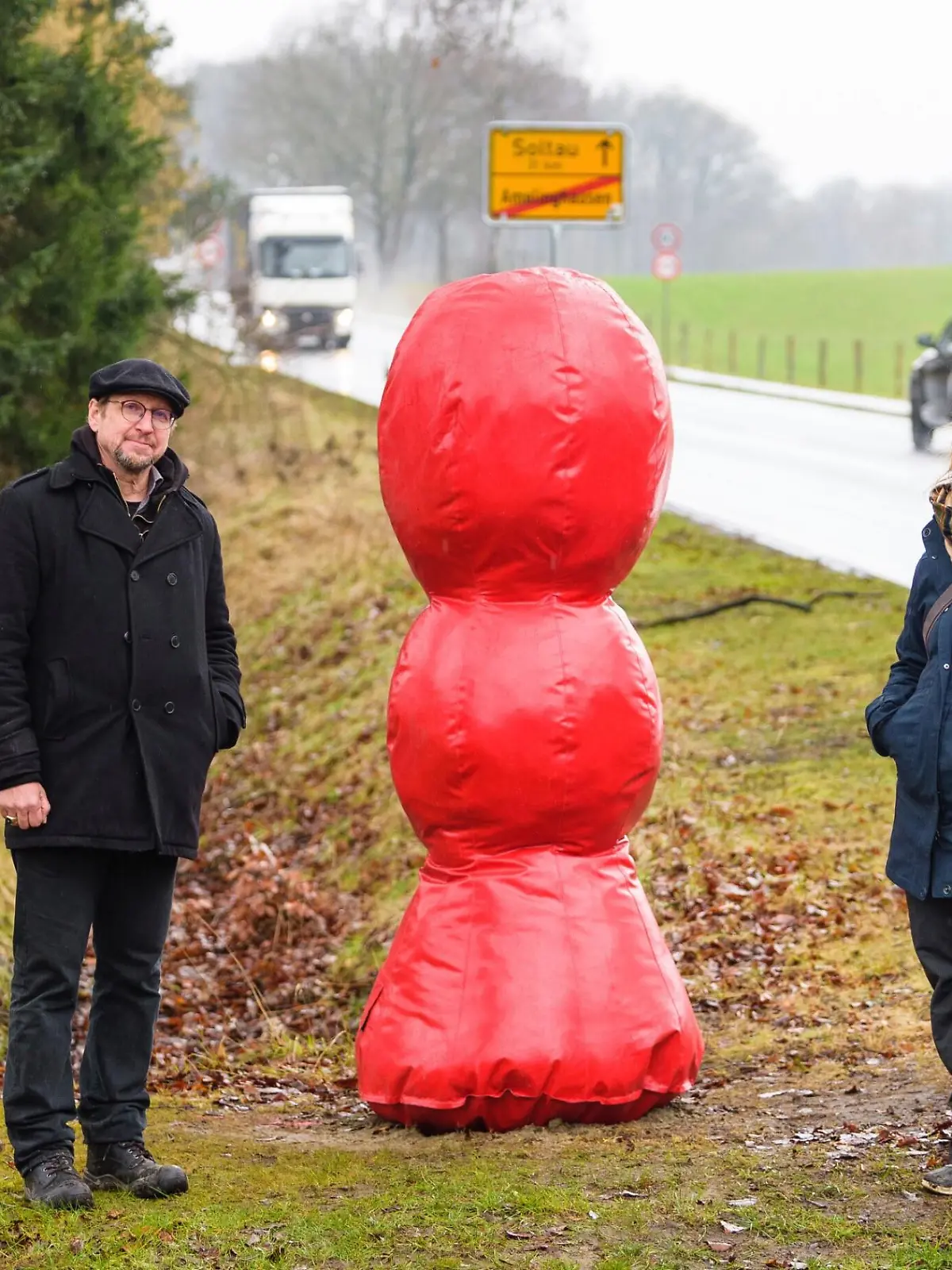 Neben einer ländlichen Straße steht eine große rote Kunstfigur zwischen zwei Menschen.