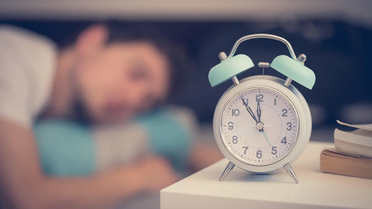White alarm clock on the bedside table. Young man sleeps in the background. || Modellfreigabe vorhanden