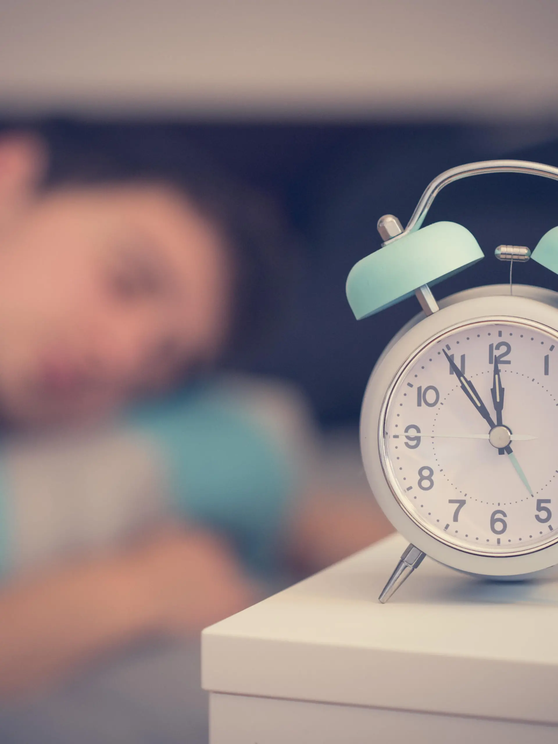 White alarm clock on the bedside table. Young man sleeps in the background. || Modellfreigabe vorhanden