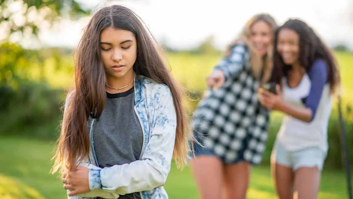 Two young teen girls point at another girl and whisper as they visibly tease her. They girls are each dressed casually and the young girl in front appears upset and shy.