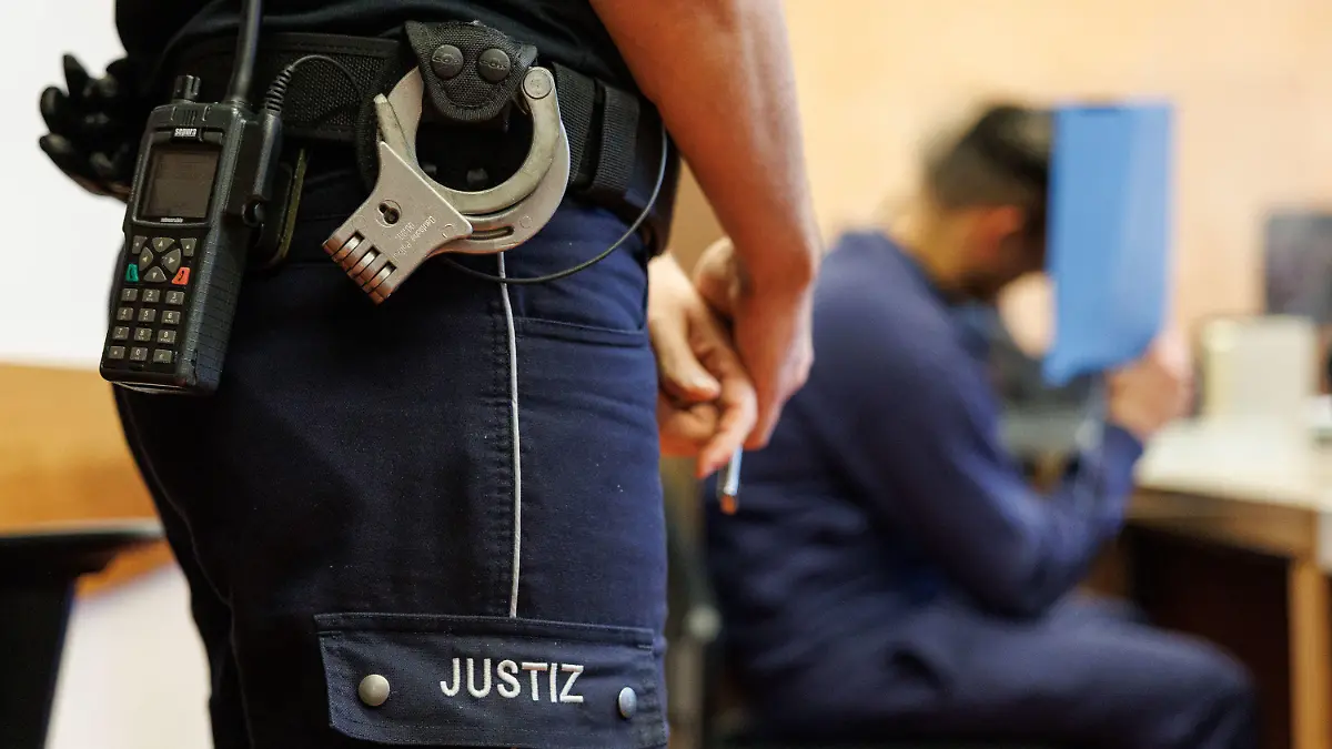 17.02.2023, Nordrhein-Westfalen, Detmold: Ein Justizbeamter, mit Funkgerät und Handschellen am Hosenbund, steht neben einem Angeklagten in einem Saal im Landgericht. Ein heute 37-Jähriger muss sich wegen Mordes und Vergewaltigung verantworten. Der Mann soll laut Anklage im Juni 2022 zuerst aus Eifersucht einen 39-Jährigen im Schlaf mit einem Axthieb getötet und anschließend seine Ex-Freundin vergewaltigt haben. Foto: Friso Gentsch/dpa +++ dpa-Bildfunk +++
