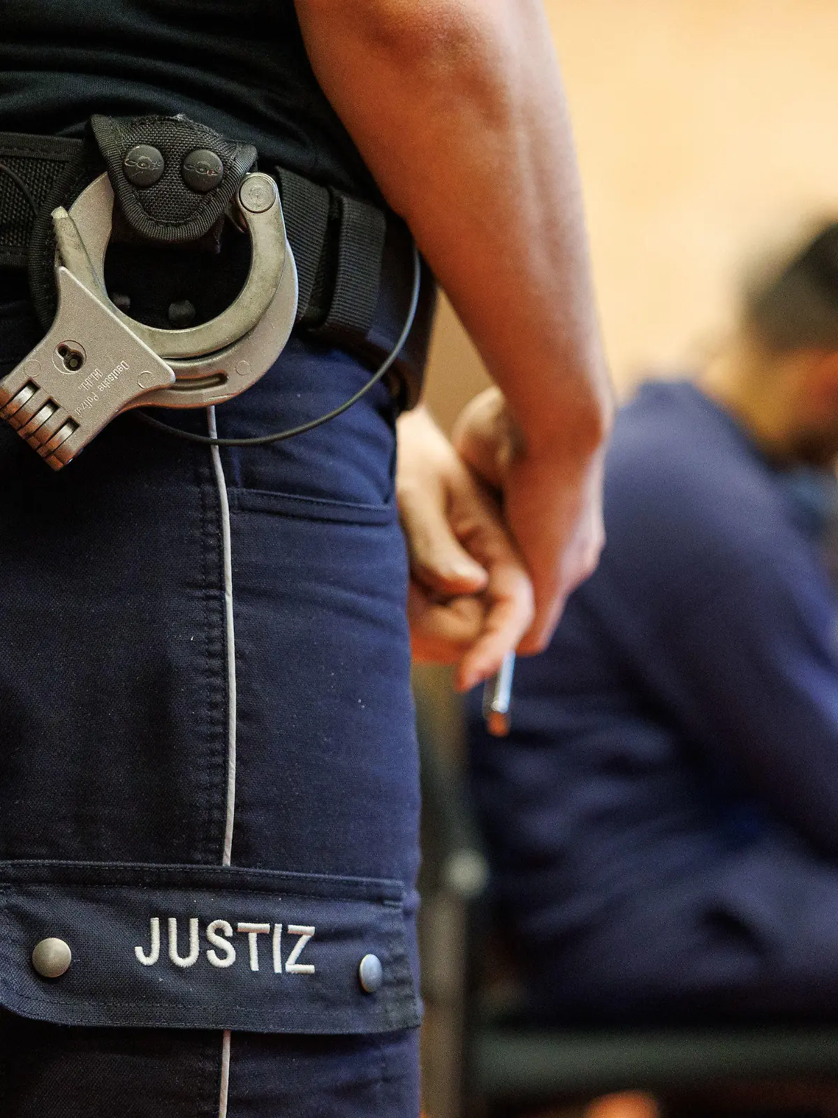 17.02.2023, Nordrhein-Westfalen, Detmold: Ein Justizbeamter, mit Funkgerät und Handschellen am Hosenbund, steht neben einem Angeklagten in einem Saal im Landgericht. Ein heute 37-Jähriger muss sich wegen Mordes und Vergewaltigung verantworten. Der Mann soll laut Anklage im Juni 2022 zuerst aus Eifersucht einen 39-Jährigen im Schlaf mit einem Axthieb getötet und anschließend seine Ex-Freundin vergewaltigt haben. Foto: Friso Gentsch/dpa +++ dpa-Bildfunk +++