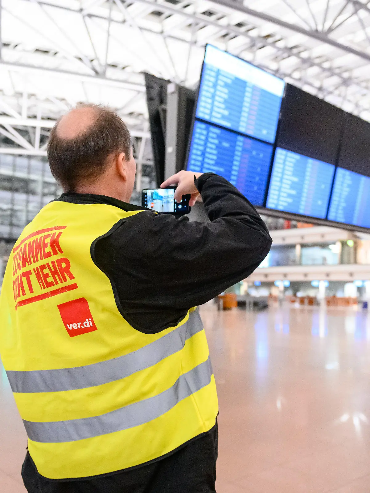 17.02.2023, Hamburg: Ein Flughafen-Mitarbeiter steht mit Streikweste im sonst menschenleeren Terminal 1 und fotografiert die Anzeigetafel auf der alle Flüge als gestrichen markiert sind. Die Gewerkschaft Verdi hatte angekündigt, in der Tarifauseinandersetzung im öffentlichen Dienst am Freitag sieben Flughäfen ganztägig lahmzulegen. Foto: Jonas Walzberg/dpa +++ dpa-Bildfunk +++