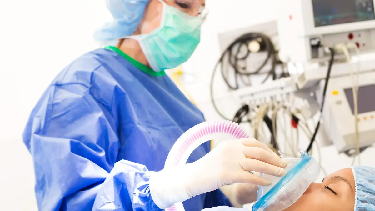 Young adult African American female patient is being sedated while laying on operating table. Senior adult Caucasian female anesthesiologist is using medical equipment to sedate patient.