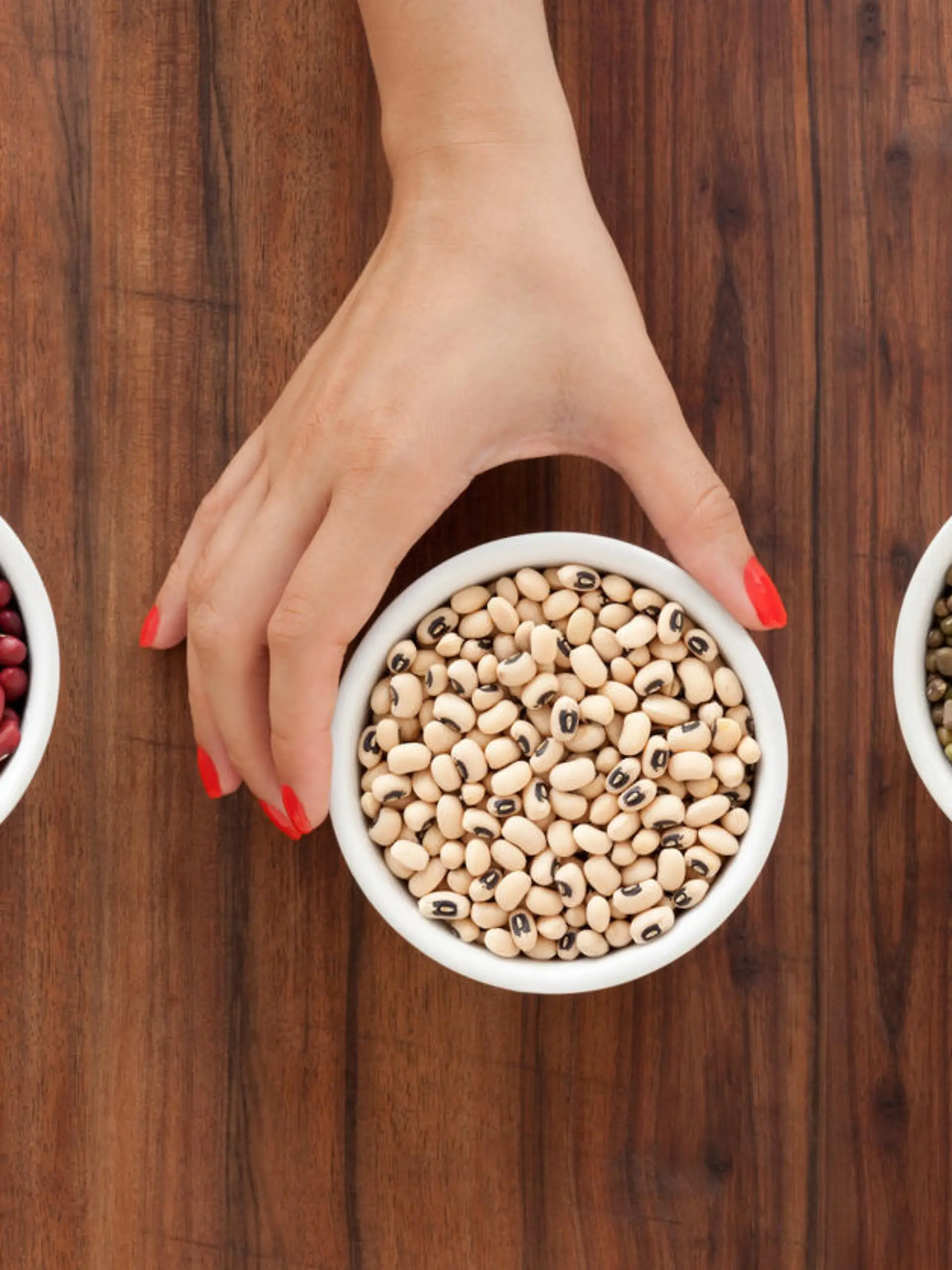 Top view of three bowls containing bean varieties and one woman hand holding the one in the middle